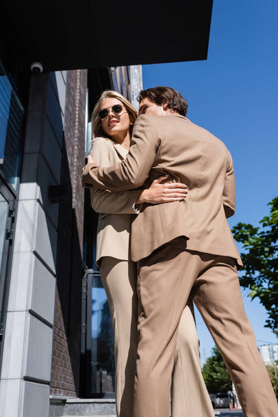 low angle view of young couple in beige suits embracing on street in city - Photo, Image