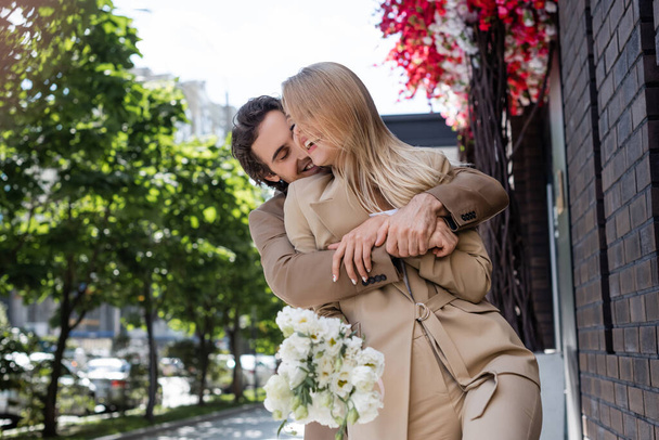 happy man embracing blonde woman holding white flowers on city street - Fotografie, Obrázek