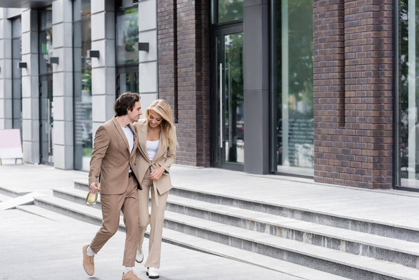 スタイリッシュなスーツ姿のカップルが建物の近くにシャンパンボトルを持って歩いていて - 写真・画像