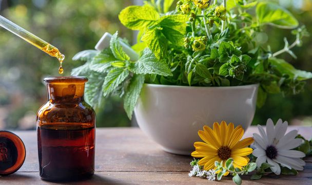 Alternative health medicine. Essential oil drop falling in a glass bottle, close up. Natural cosmetics, serum and vitamins. Fresh herbs in a mortar on a wooden table - Φωτογραφία, εικόνα