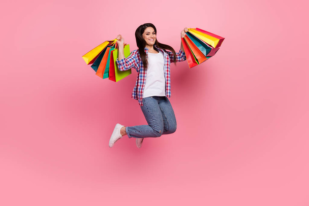 Full size photo of mature brunette lady hold bags jump wear shirt jeans sneakers isolated on pink background - Φωτογραφία, εικόνα