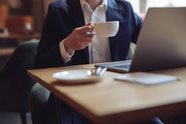 Cup of coffee in male hand of caucasian elegant businessman. Working at laptop in cafe. - Foto, Imagen