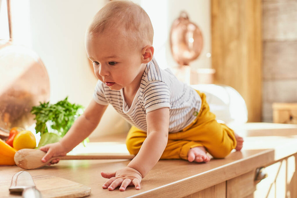 A cute baby boy, an infant, wearing yellow pants and a striped T-shirt, sits on the kitchen table. A small child cooks with his mother in the kitchen, crawls around the table, explores the products. - Фото, изображение