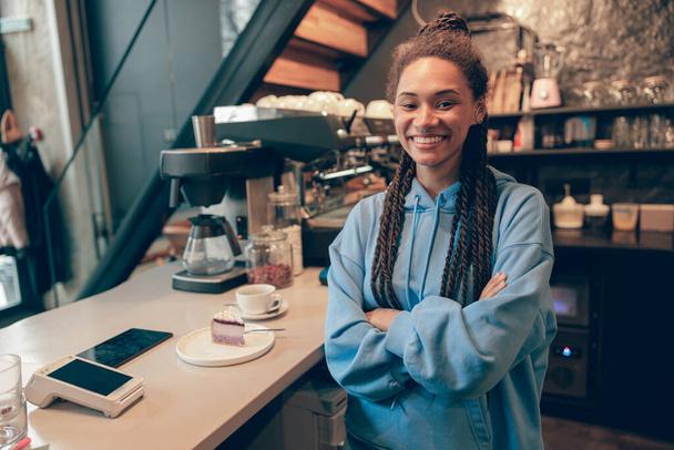 Όμορφη νεαρή ευτυχισμένη κομψή γυναίκα barista στέκεται και χαμογελά στον πάγκο στην καφετέρια. - Φωτογραφία, εικόνα