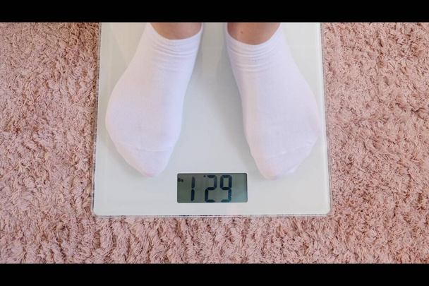 Vrouw benen in witte sokken staan op digitale schalen om gewicht te controleren op de vloer in de kamer. - Foto, afbeelding