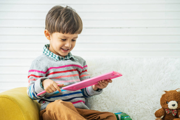 Guapo chico de raza mixta, feliz diversión y sonrisa usando lápiz de tablero de juguete para dibujar, niño disfrutando desarrollando actividades en casa, jugando en el sofá. - Foto, Imagen