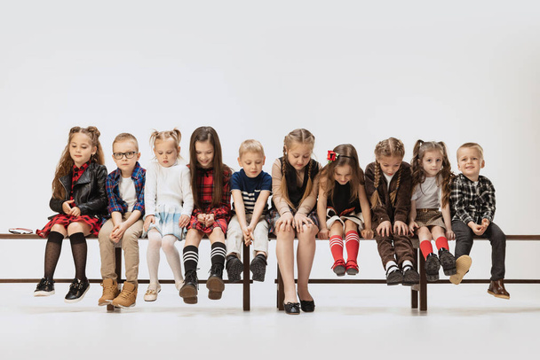 Group of school and preschool age children, girls and boys sitting on banch isolated on grey studio background. Beauty, kids fashion, education, happy childhood concept. - Fotó, kép