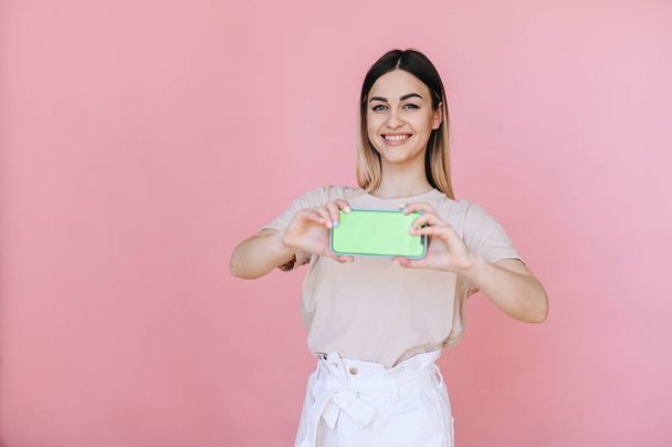 Uma menina com um sorriso segura um telefone inteligente horizontalmente com uma tela verde em um fundo rosa. Lugar para publicidade ou mensagem. slogan - Foto, Imagem
