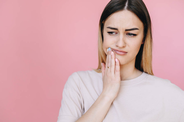 The girl suffers from a toothache, frowns her face and looks away on a pink background. Dentistry concept. Advertising on a banner - Photo, image