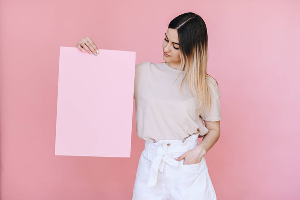Девушка держит в правой руке лист розовой бумаги, а правая рука в кармане на розовом фоне. - Фото, изображение