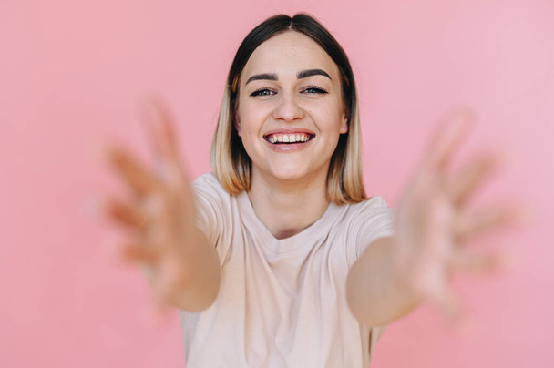 A young woman stretches her arms forward while hugging on a pink background. The hands in the foreground are out of focus. - Фото, изображение