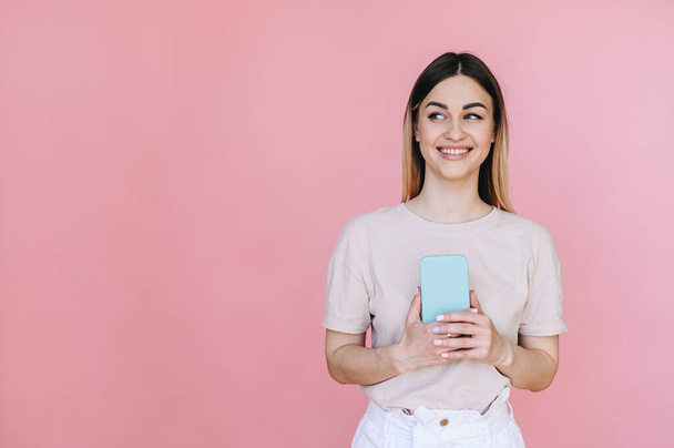 Een meisje met een mysterieuze uitdrukking kijkt weg en houdt een telefoon in haar handen op een roze achtergrond. Links is een vrije ruimte voor tekst. - Foto, afbeelding