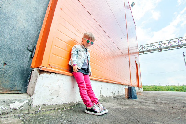 Little stylish girl in sunglasses with craft bags on shopping trip on an orange background. Shopaholic dances against the wall of the commercial center. Girl happy shopping on Black Friday. Copy space - Zdjęcie, obraz