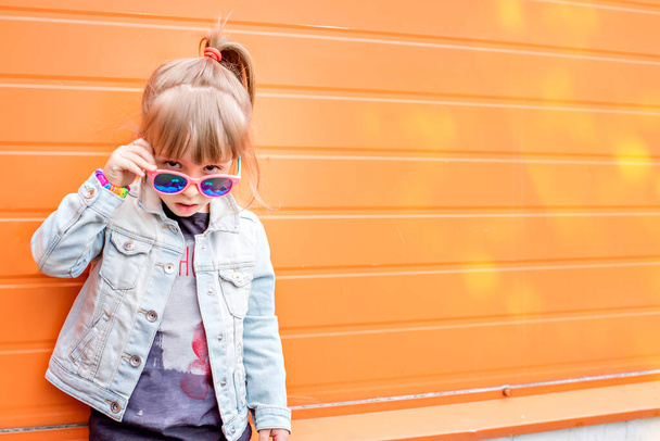 Emotional close up portrait of Little stylish girl in sunglasses on an orange background.The girl in a denim jacket grimaces, has fun, shows her tongue. Copy space - Photo, image