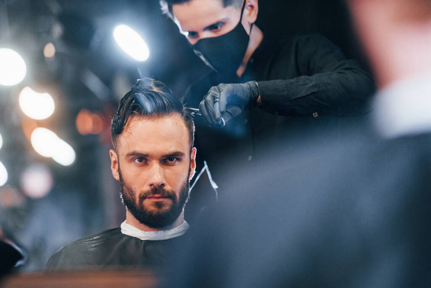 Front view of young bearded man that sitting and getting haircut in barber shop by guy in black protective mask. - Valokuva, kuva