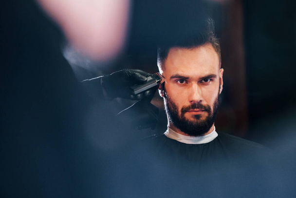 Young bearded man sitting and getting haircut in barber shop. - Foto, Imagem