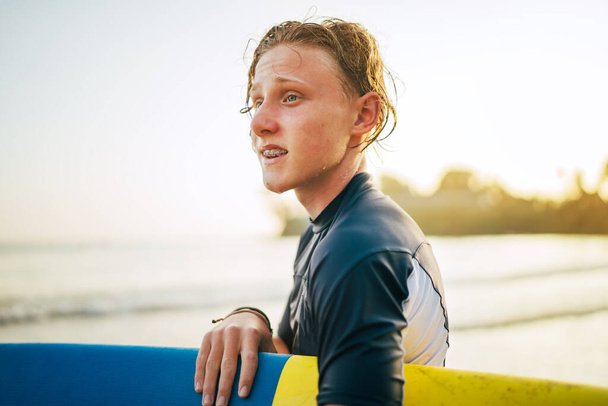 Portrait of a teen boy with Dental braces and wet hair with a surfboard goes for surfing. He is smiling and walking into the water. Happy boyhood and active vacation time concept. - Photo, image