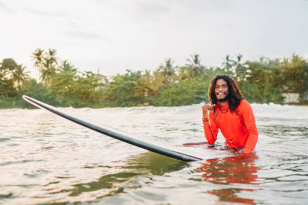 Black long-haired teen man showing shaka sign floating on long surfboard, waiting wave ready surfing with palms litted sunset rays. Extreme water sports or traveling to exotic countries concept - Photo, image