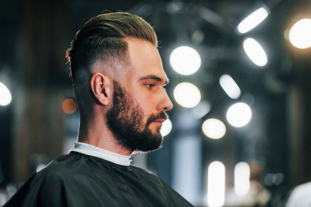 Young bearded man sitting in barber shop and waiting for stylist. - Photo, image