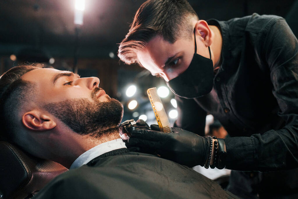 Молодой человек со стильной прической сидит и сбривает бороду парнем в черной защитной маске в парикмахерской. - Фото, изображение