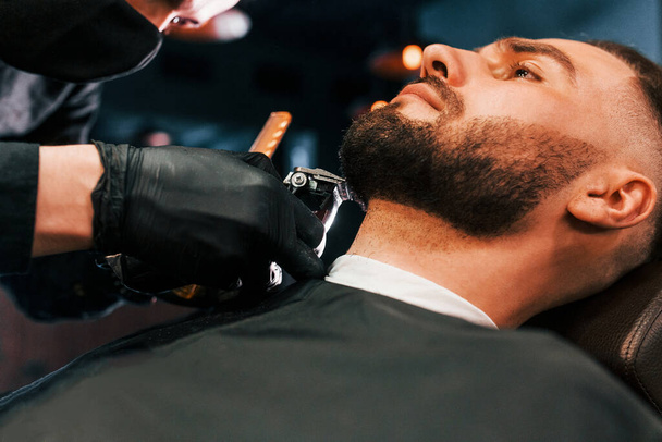 Young man with stylish hairstyle sitting and getting his beard shaved in barber shop. - Foto, Imagem