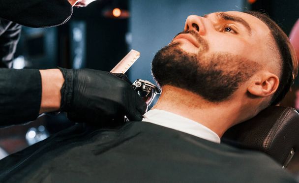 Young man with stylish hairstyle sitting and getting his beard shaved in barber shop. - Foto, Bild