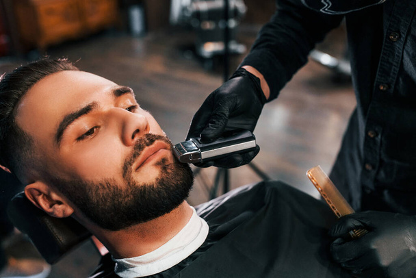 Молодий чоловік зі стильною зачіскою сидить і голиться борода в перукарні
. - Фото, зображення