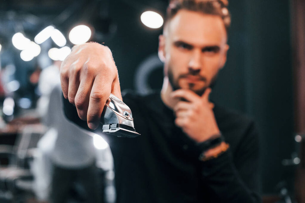 Young bearded man standing in barber shop and holding clipper. - Foto, Imagem