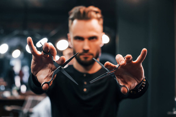 Young bearded man standing in barber shop and holding scissors. - Photo, image