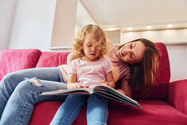Reading big book. Young mother with her little daughter in casual clothes together indoors at home. - Photo, Image