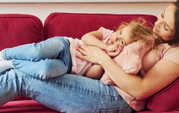 Lying down on red sofa. Young mother with her little daughter in casual clothes together indoors at home. - Foto, imagen