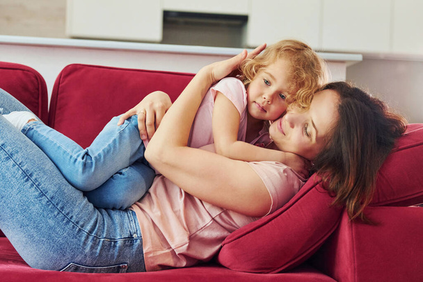 Lying down on red sofa. Young mother with her little daughter in casual clothes together indoors at home. - 写真・画像
