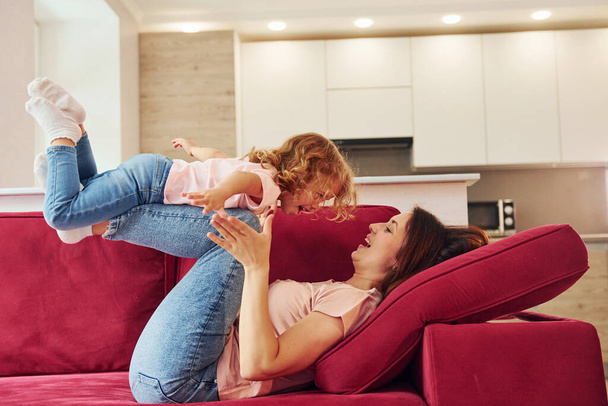 Having fun on red sofa. Young mother with her little daughter in casual clothes together indoors at home. - Foto, Bild