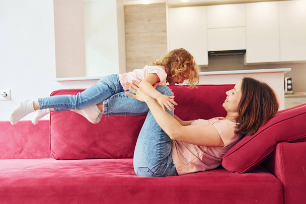 Having fun on red sofa. Young mother with her little daughter in casual clothes together indoors at home. - Fotografie, Obrázek