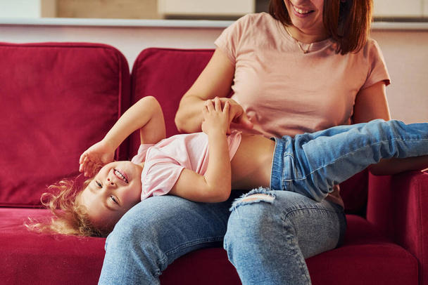 Lying down on red sofa. Young mother with her little daughter in casual clothes together indoors at home. - Fotoğraf, Görsel