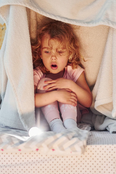 Sitting under blanket. Cute little girl in casual clothes is indoors at home at daytime. - Foto, Imagen