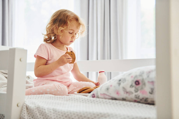 Op het bed zitten, koekjes eten en melk drinken. Schattig klein meisje in casual kleding is binnen in huis overdag. - Foto, afbeelding