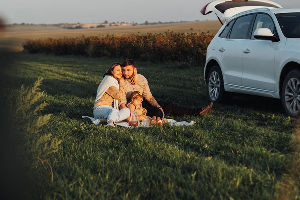 Happy Young Family Enjoying Road Trip on SUV Car, Mom Dad and their Baby Daughter Having Picnic on Weekend Outdoors at Sunset - Zdjęcie, obraz