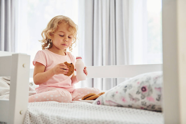 Sitting on the bed, eating cookies and drinking milk. Cute little girl in casual clothes is indoors at home at daytime. - Foto, Imagen