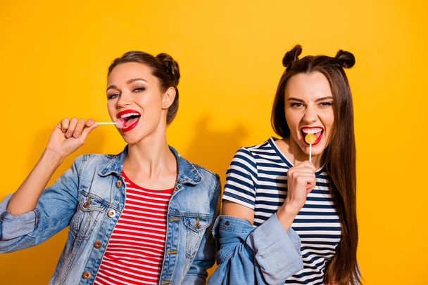 Portrait of two excited carefree pretty girls enjoy lollipop stick isolated on yellow color background - Фото, зображення