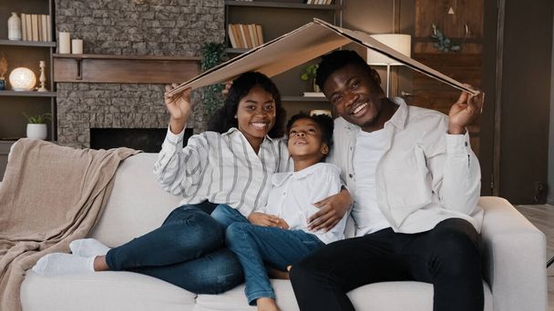 African couple sitting on couch under cardboard roof smiling looking at camera active happy daughter girl running sits on sofa. Relocation day to new home moving to own apartment homeowner portrait - Photo, image