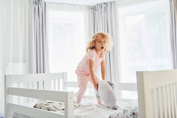 Playing with pillow by throwing it. Cute little girl in casual clothes is indoors at home at daytime. - Fotó, kép