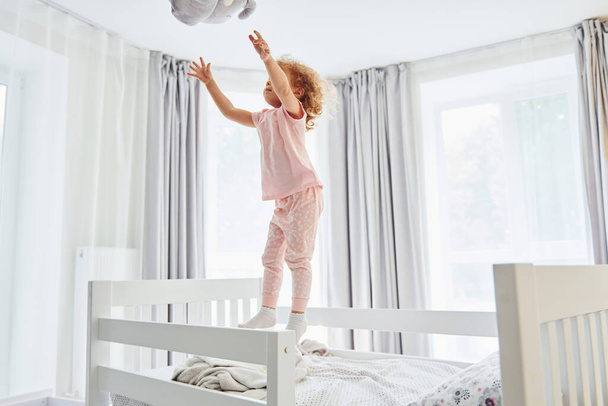 Playing with pillow by throwing it. Cute little girl in casual clothes is indoors at home at daytime. - Фото, изображение