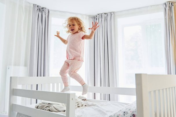 Jumping on the bed. Cute little girl in casual clothes is indoors at home at daytime. - Photo, image