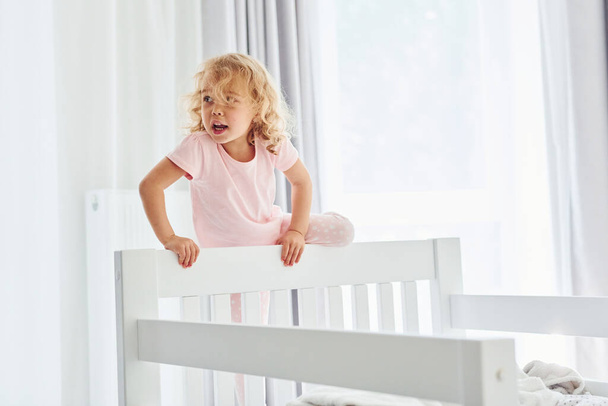 Standing on the bed. Cute little girl in casual clothes is indoors at home at daytime. - Foto, Imagen