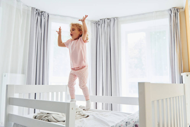 Jumping on the bed. Cute little girl in casual clothes is indoors at home at daytime. - Fotografie, Obrázek