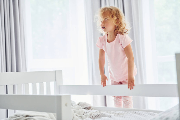 Standing on the bed. Cute little girl in casual clothes is indoors at home at daytime. - Foto, afbeelding