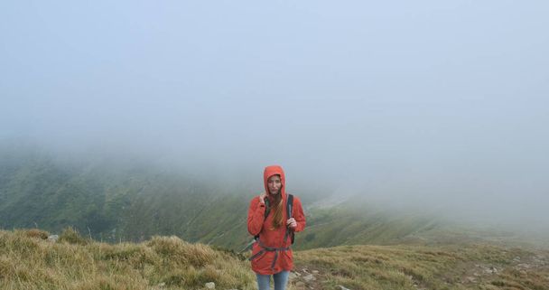 Woman look into the camera, standing on the edge of the mountain in a gray cloud. Movement of fog on the background. Stormy weather at the summit. Medium shot, spring summer, Carpathians, Ukraine. - Foto, Imagem