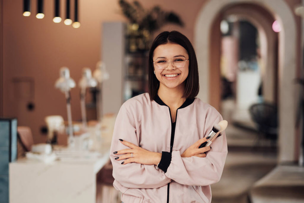 Portrait of Young Cheerful Female Make Up Artist Standing in the Beauty Salon, Caucasian Woman in Glasses with Folded Arms Holding Brushes - Foto, Bild