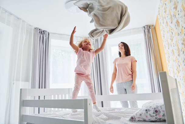 Having fun by throwing a pillow. Young mother with her little daughter in casual clothes together indoors at home. - Foto, Imagem